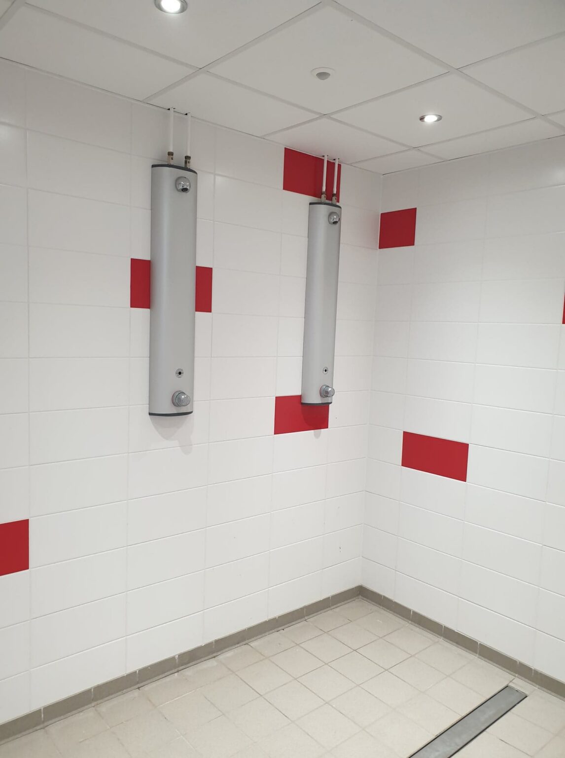 Les douches d'un vestiaire : deux colonnes de douche sur un carrelage blanc semé de carreaux rouges, avec une rigole d'évacuation au sol.