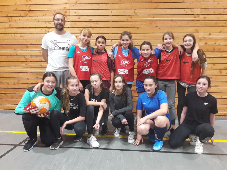 L'équipe féminine dans une salle de sport : sept joueuses debout en chasubles rouges, l'entraîneur à gauche, et six accroupies devant, dont la gardienne qui tient le ballon ; les noms sont donnés dans le texte voisin.