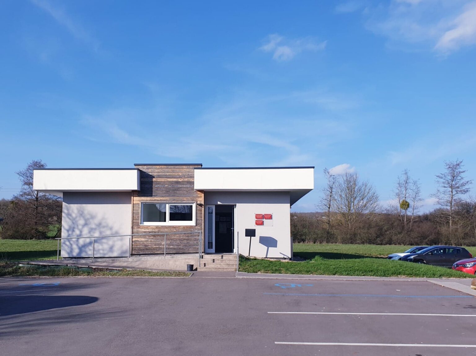 Un bâtiment moderne de plain-pied, blanc et bardé de bois, avec une porte vitrée, des boîtes aux lettres rouges et une rampe d'accès ; devant, un parking avec des places réservées.