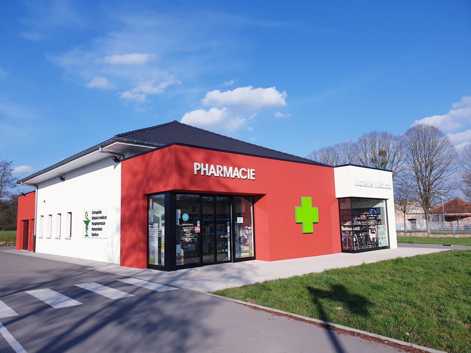 La pharmacie : un bâtiment moderne rouge et blanc portant l'inscription « Pharmacie », une croix verte lumineuse, une vitrine « Matériel médical » et un panneau énumérant orthopédie, parapharmacie, herboristerie et nutrition.