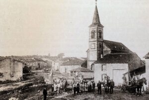 Vue ancienne du village : l'église et son clocher pointu dominent une rue en pente bordée de maisons basses ; au premier plan, sur la terre battue, un groupe de villageois, des enfants et deux chevaux attelés.