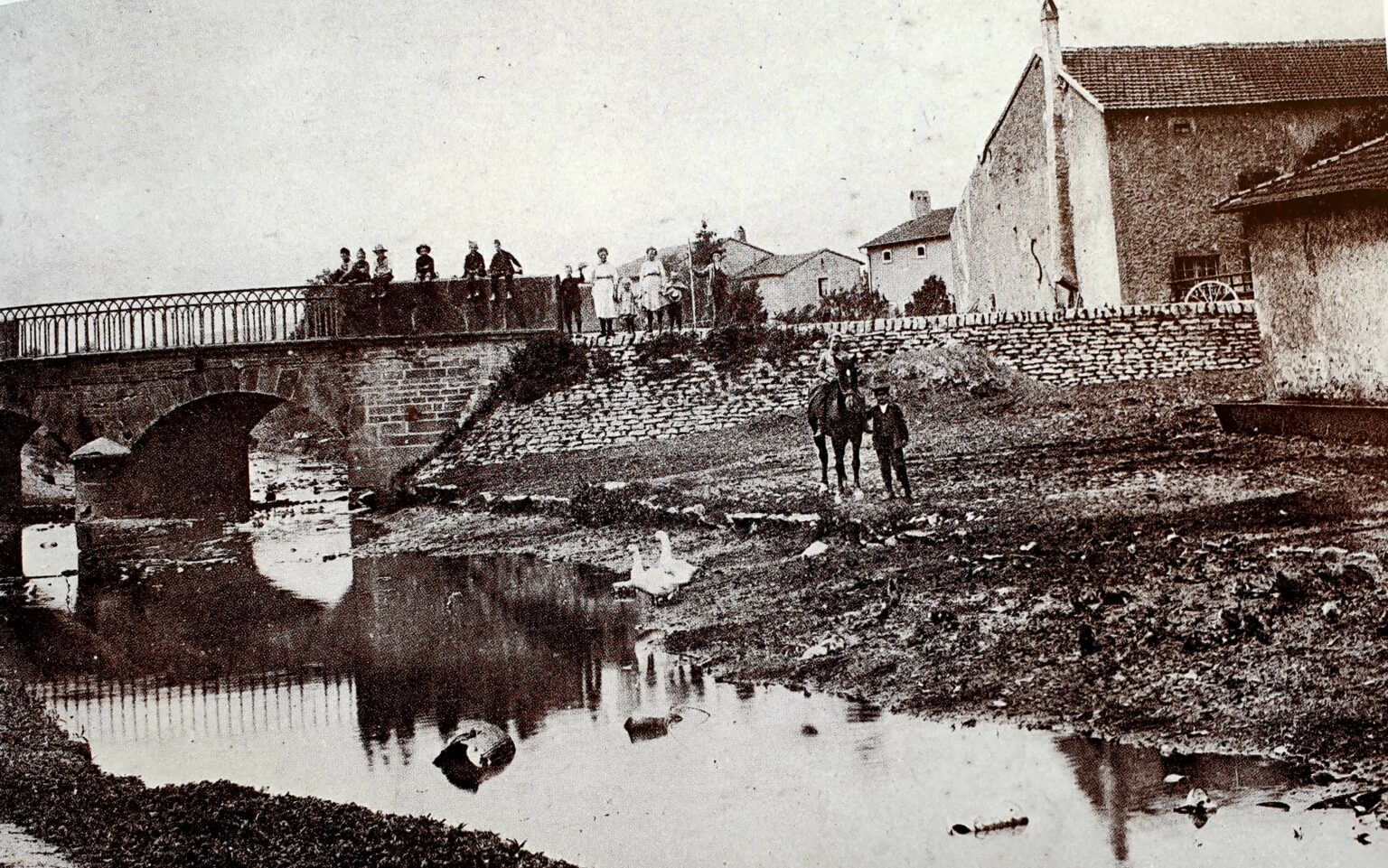 Un pont de pierre à garde-corps de fer enjambe le ruisseau. Des enfants sont assis sur le parapet, des femmes se tiennent debout à côté, et un homme mène un cheval sur la berge pavée, devant les maisons du village.