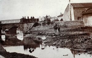Un pont de pierre à garde-corps de fer enjambe le ruisseau. Des enfants sont assis sur le parapet, des femmes se tiennent debout à côté, et un homme mène un cheval sur la berge pavée, devant les maisons du village.