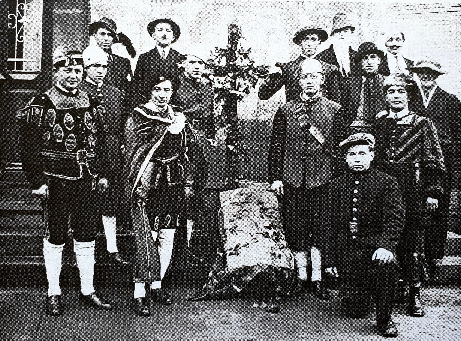 Une quinzaine de villageois en costume de scène — tuniques, capes, chapeaux à plume, culottes courtes et bas blancs — posent sur les marches d'une maison, autour d'un décor de feuillage.