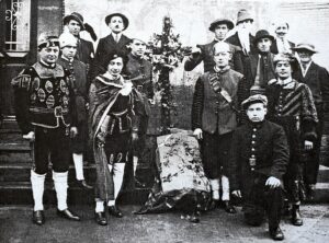 Une quinzaine de villageois en costume de scène — tuniques, capes, chapeaux à plume, culottes courtes et bas blancs — posent sur les marches d'une maison, autour d'un décor de feuillage.