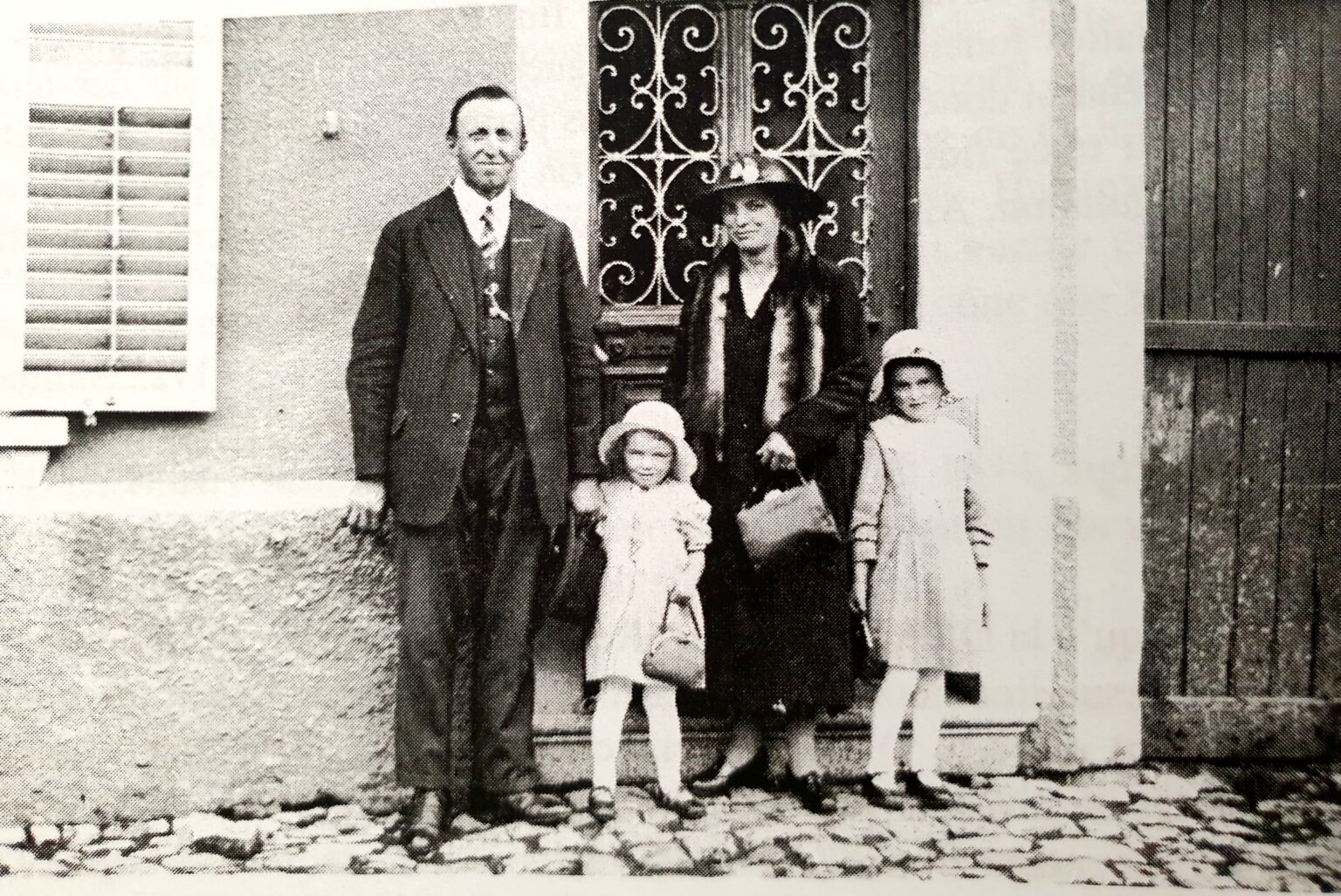 Un homme en costume, une femme en manteau et chapeau cloche, et deux fillettes en manteau clair et béguin posent sur les pavés, devant une porte de maison à grille de fer forgé.