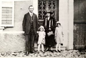 Un homme en costume, une femme en manteau et chapeau cloche, et deux fillettes en manteau clair et béguin posent sur les pavés, devant une porte de maison à grille de fer forgé.
