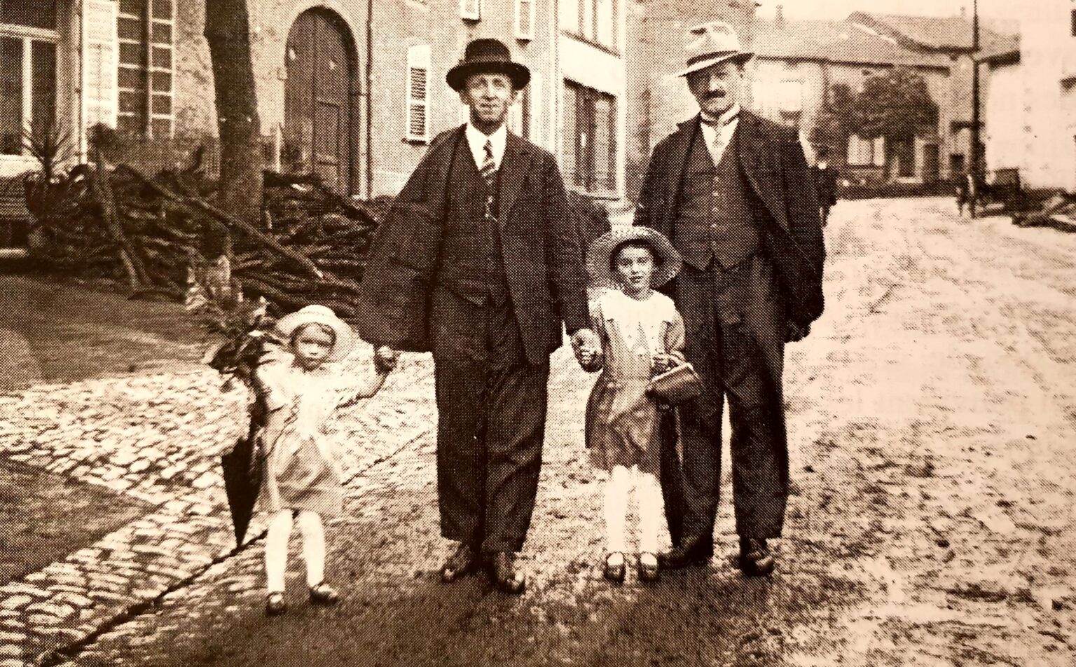 Deux hommes en costume et chapeau tiennent par la main deux fillettes en robe claire et béguin, sur une rue pavée du village ; derrière eux, un tas de fagots contre une maison.