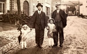 Deux hommes en costume et chapeau tiennent par la main deux fillettes en robe claire et béguin, sur une rue pavée du village ; derrière eux, un tas de fagots contre une maison.