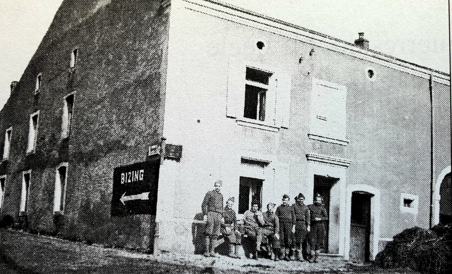 À l'angle de deux maisons, un panneau noir indiquant « BIZING » avec une flèche blanche ; une dizaine de soldats en uniforme sont rassemblés le long de la façade claire.