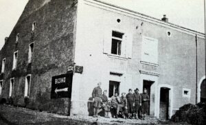 À l'angle de deux maisons, un panneau noir indiquant « BIZING » avec une flèche blanche ; une dizaine de soldats en uniforme sont rassemblés le long de la façade claire.