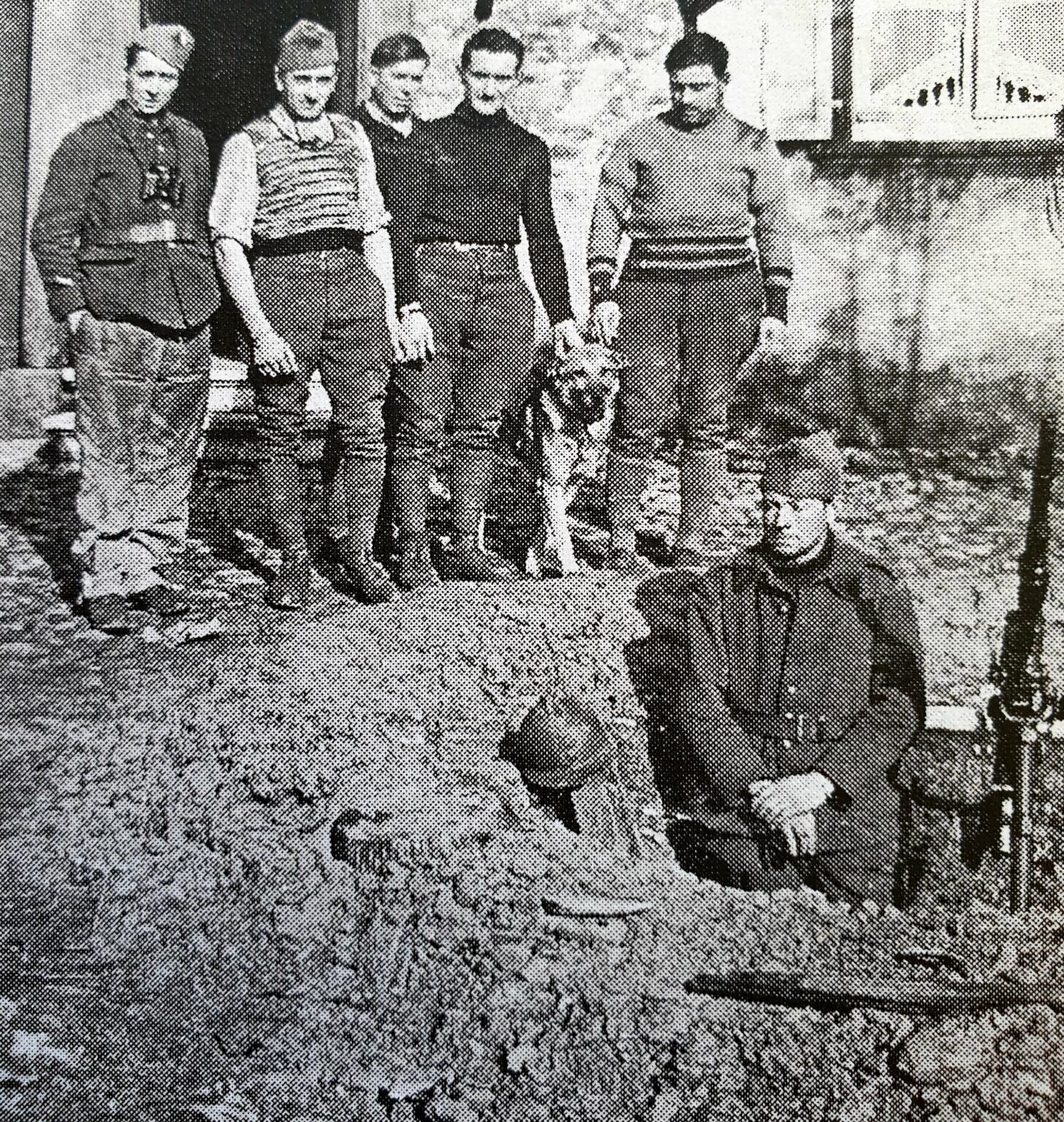 Cinq jeunes hommes debout au bord d'un talus de terre et un sixième agenouillé à droite, en tenue militaire, devant une maison du village.