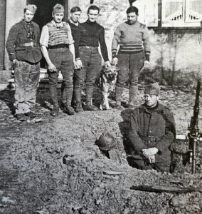 Cinq jeunes hommes debout au bord d'un talus de terre et un sixième agenouillé à droite, en tenue militaire, devant une maison du village.
