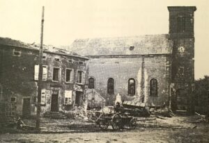 L'église après la guerre : la tour du clocher est réduite à un moignon sans flèche, le mur de la nef est marqué d'impacts et la maison voisine est éventrée ; une charrette attelée stationne sur la place.