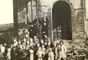 Une trentaine d'enfants rassemblés devant le portail de l'église ; au-dessus d'eux, des hommes sont montés sur une échelle appuyée contre la façade, et des gravats sont entassés à droite.