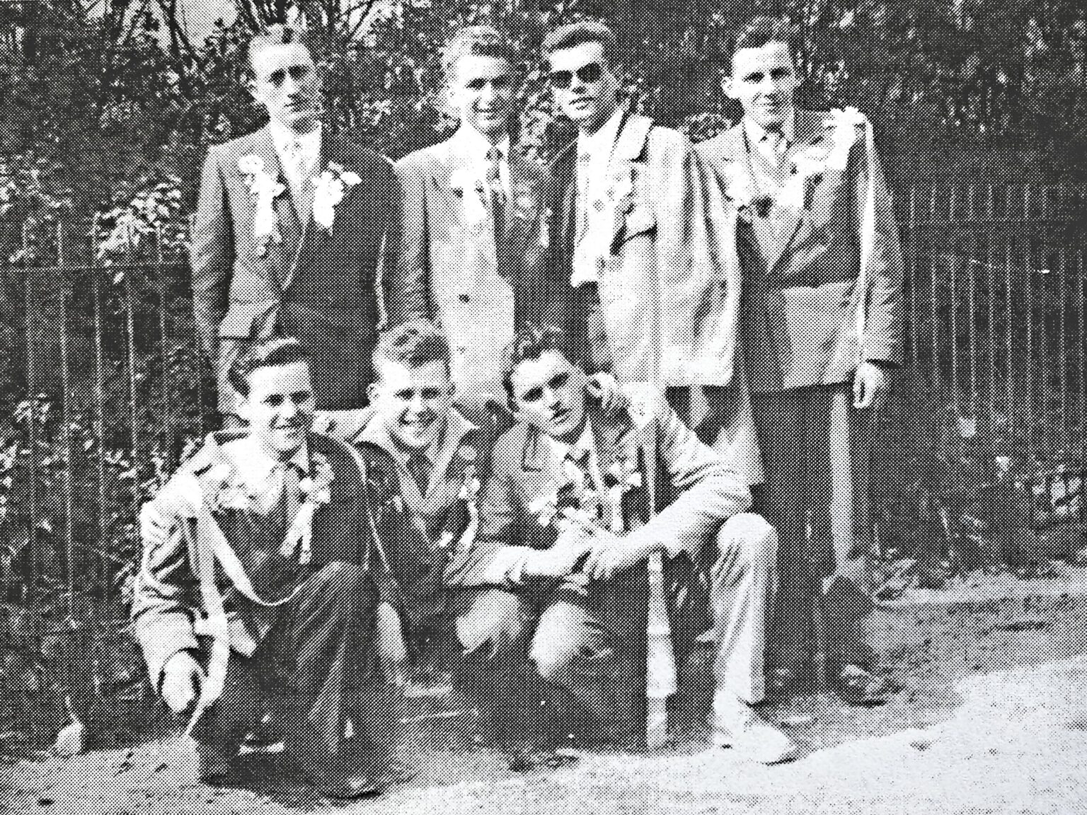 Huit jeunes hommes en veste et chemise ouverte, quatre debout et quatre accroupis au premier rang, posent devant un bosquet ; plusieurs portent des lunettes de soleil.