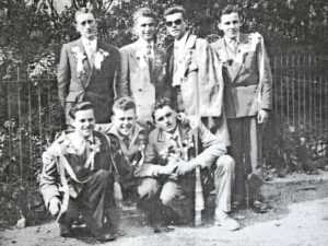 Huit jeunes hommes en veste et chemise ouverte, quatre debout et quatre accroupis au premier rang, posent devant un bosquet ; plusieurs portent des lunettes de soleil.