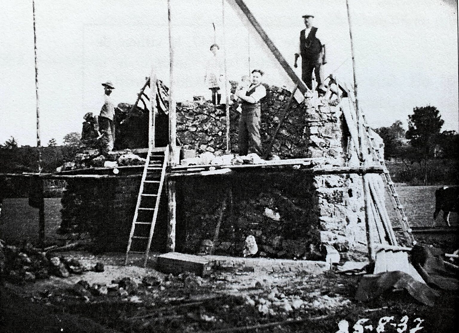 Chantier de la grotte : des hommes travaillent sur un échafaudage de planches et deux échelles, au-dessus d'un mur de pierres en cours de montage ; la date « 25-8-37 » est inscrite en bas de la photographie.