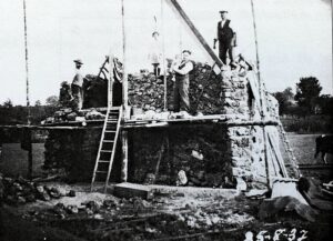 Chantier de la grotte : des hommes travaillent sur un échafaudage de planches et deux échelles, au-dessus d'un mur de pierres en cours de montage ; la date « 25-8-37 » est inscrite en bas de la photographie.