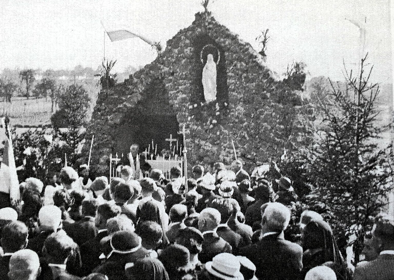 Une foule compacte, chapeaux, bannières et croix de procession levées, fait face à la grotte couverte de lierre, dans la niche de laquelle se détache une statue blanche de la Vierge.