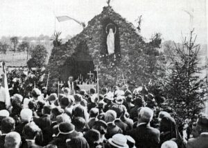 Une foule compacte, chapeaux, bannières et croix de procession levées, fait face à la grotte couverte de lierre, dans la niche de laquelle se détache une statue blanche de la Vierge.