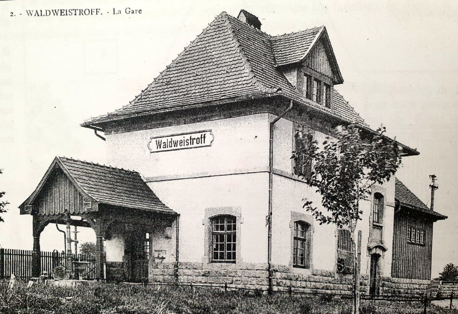 Carte postale de la gare de Waldweistroff : un bâtiment à large toit de tuiles portant le panneau « Waldweistroff », avec un auvent de bois sur la gauche et une clôture devant.