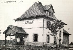 Carte postale de la gare de Waldweistroff : un bâtiment à large toit de tuiles portant le panneau « Waldweistroff », avec un auvent de bois sur la gauche et une clôture devant.