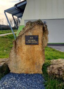 Un gros bloc de pierre calcaire porte une plaque sombre aux lettres dorées : « Place Saint Gervais les Trois Clochers ». Il est posé sur l'herbe, devant un bâtiment moderne à auvent métallique.