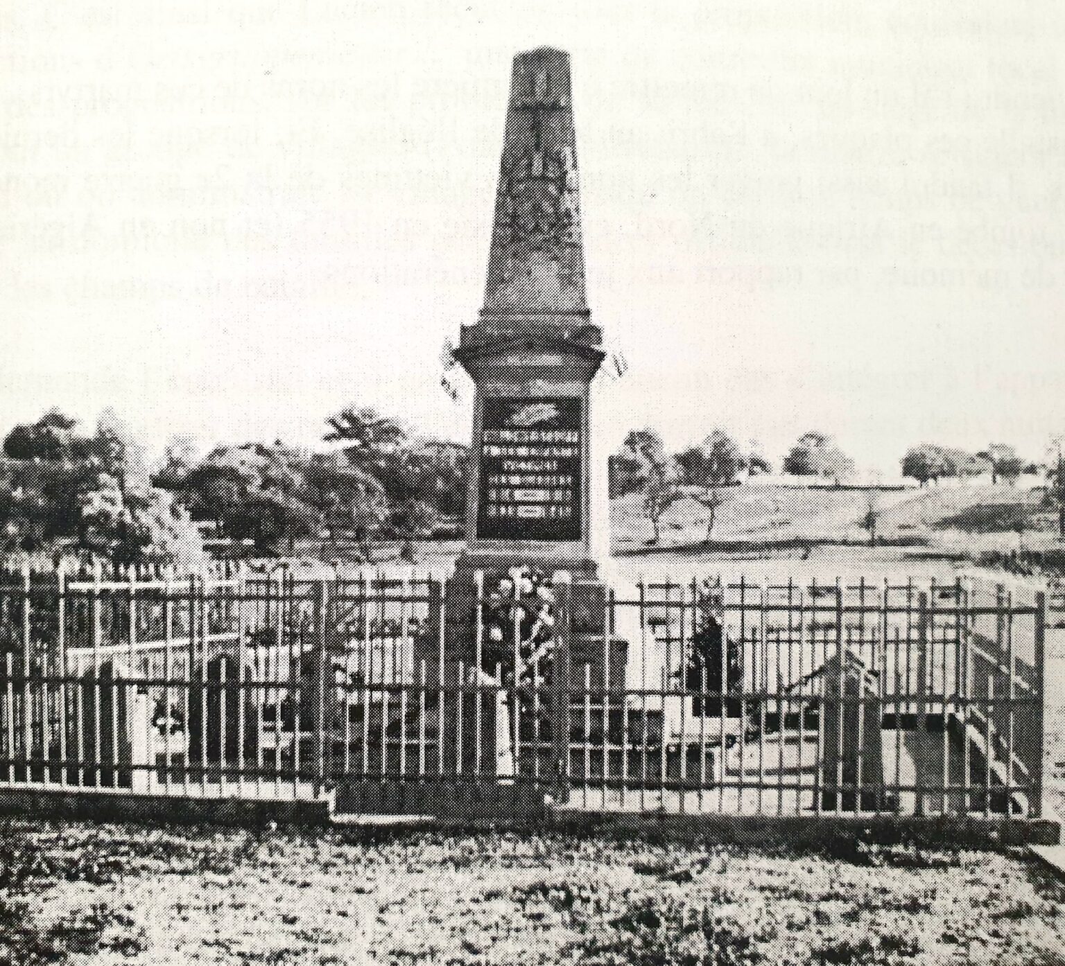 Le monument aux morts : un haut obélisque de pierre dressé sur un socle gravé, derrière une grille de fer, au milieu des tombes et des croix du cimetière.