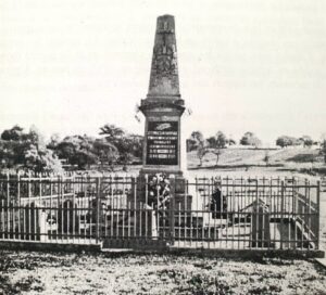 Le monument aux morts : un haut obélisque de pierre dressé sur un socle gravé, derrière une grille de fer, au milieu des tombes et des croix du cimetière.