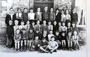 Photographie de classe : une quarantaine de garçons posent sur trois rangs devant un mur de pierre et une porte de bois ; l'un d'eux, assis au sol, tient une ardoise portant un numéro.