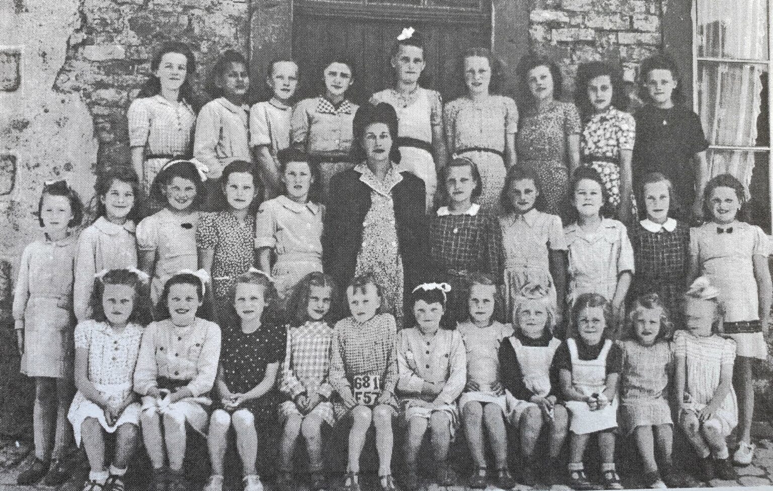 Photographie de classe : une quarantaine de filles posent sur trois rangs, l'institutrice au milieu du groupe ; une élève assise tient une ardoise portant un numéro.