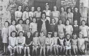 Photographie de classe : une quarantaine de filles posent sur trois rangs, l'institutrice au milieu du groupe ; une élève assise tient une ardoise portant un numéro.