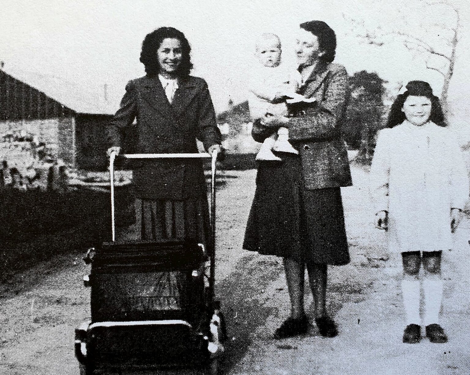 Deux femmes sur un chemin, l'une poussant un landau, l'autre portant un bébé dans les bras ; à droite, une fillette en robe claire lève la main.