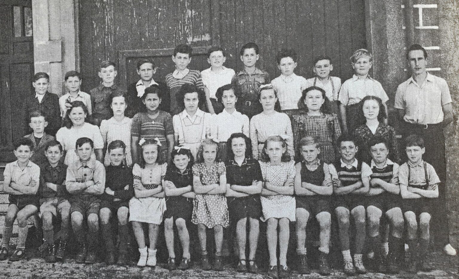 Photographie de classe mixte : environ trente-cinq garçons et filles posent sur trois rangs, les plus jeunes assis au premier plan, bras croisés.