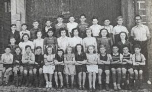 Photographie de classe mixte : environ trente-cinq garçons et filles posent sur trois rangs, les plus jeunes assis au premier plan, bras croisés.
