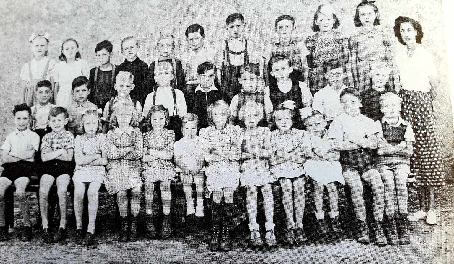 Photographie de classe : une trentaine de jeunes enfants, garçons et filles, posent sur trois rangs, l'institutrice debout à droite du groupe.