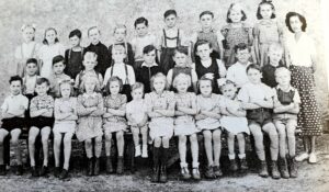 Photographie de classe : une trentaine de jeunes enfants, garçons et filles, posent sur trois rangs, l'institutrice debout à droite du groupe.