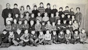 Photographie de classe en noir et blanc : une cinquantaine d'enfants en quatre rangs — assis en tailleur au premier plan, accroupis, puis debout — devant un mur clair ; un instituteur se tient à gauche, une institutrice à droite, et une ardoise posée au sol en bas à gauche porte une inscription à la craie.