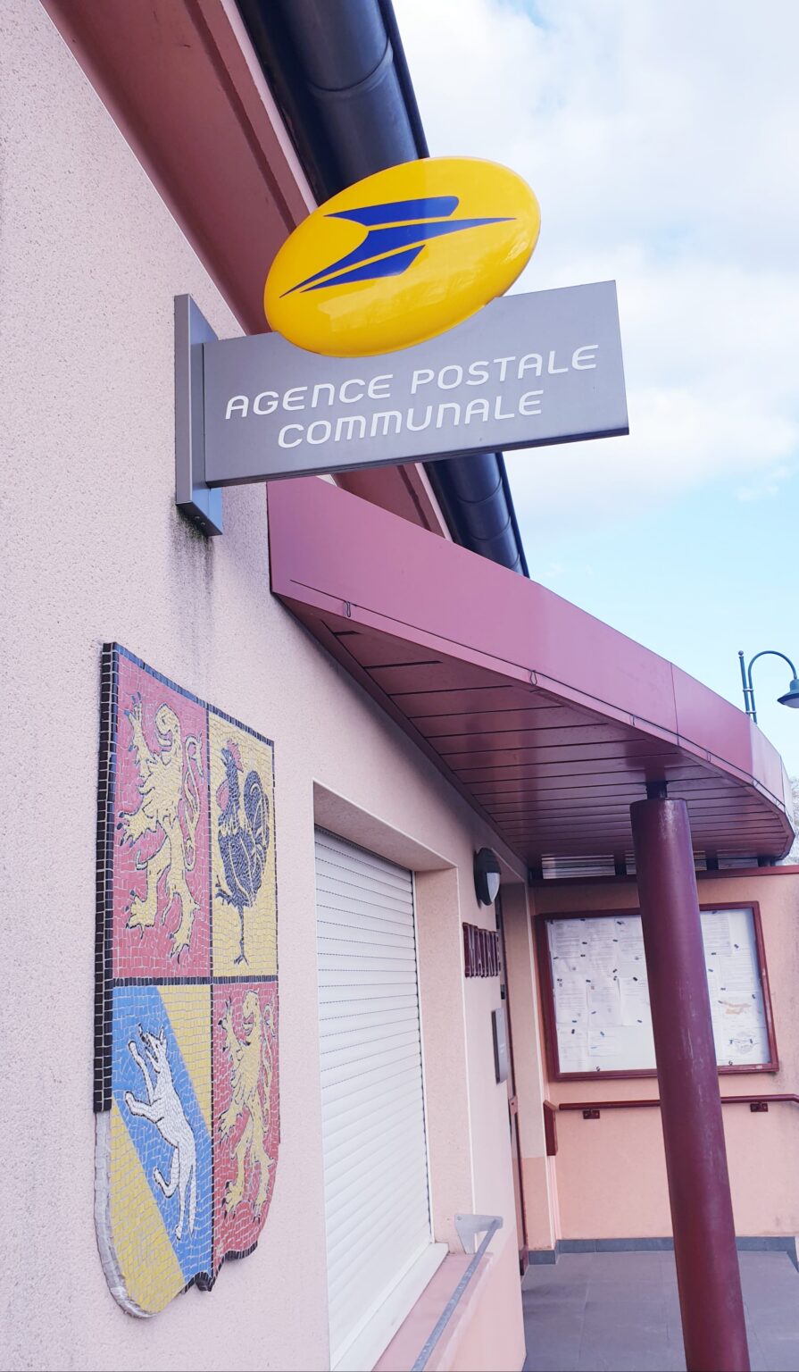L'enseigne jaune de La Poste et une plaque grise « Agence postale communale » au-dessus de l'entrée ; sur le mur, le blason de la commune en mosaïque.