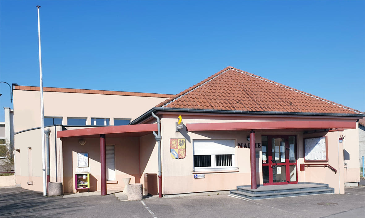 La mairie : un bâtiment beige à toit de tuiles rouges, l'inscription « Mairie » au-dessus d'une porte vitrée rouge, le blason de la commune sur la façade, des panneaux d'affichage de part et d'autre et une rampe le long du perron.