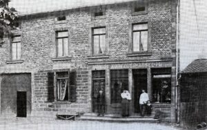 Une maison de pierre à devanture de boutique, l'enseigne courant au-dessus des vitrines ; un homme en tablier blanc de boucher tient un enfant par la main, à côté d'une femme et d'un autre homme sur le seuil.