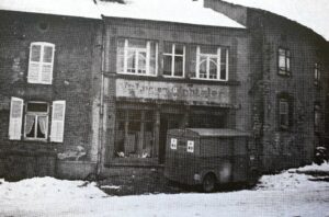Une devanture de magasin sous la neige, l'enseigne effacée au-dessus des vitrines ; une camionnette est garée devant, et la rue est couverte de neige.