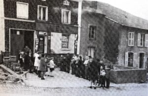 Un groupe d'habitants, surtout des femmes et des enfants, rassemblé devant l'entrée d'un magasin, au bord d'une rue bordée de maisons.