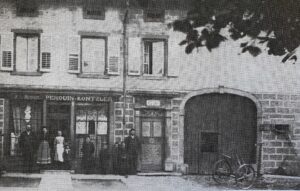 Un magasin dont l'enseigne porte « Michel Perquin-Kontzler » ; quelques personnes posent dans l'embrasure de la porte, et une grande porte cochère en plein cintre ouvre à droite.