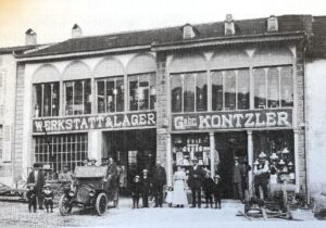 Une large devanture à arcades portant les enseignes « Werkstatt & Lager » et « Gebr. Kontzler » ; une vingtaine de personnes posent devant les vitrines garnies, à côté d'une automobile ancienne.