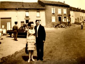 Un couple pose au milieu de la rue : une femme en robe claire à volants, un sac à la main, et un homme en costume sombre ; derrière eux, une pompe à essence, une voiture des années soixante et les maisons du village.