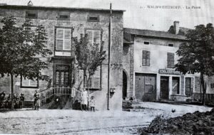 Carte postale de la poste : un bâtiment de pierre auquel on accède par un perron à rampe, encadré d'arbres ; à droite une boutique à enseigne, et quelques habitants sur la place de terre.