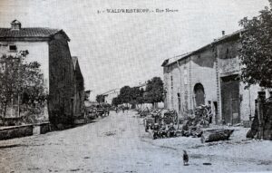 Carte postale de la « Rue Neuve » : une large rue de terre bordée à gauche d'une grande maison à porte de grange, à droite d'un alignement de maisons ; une poule picore au premier plan.