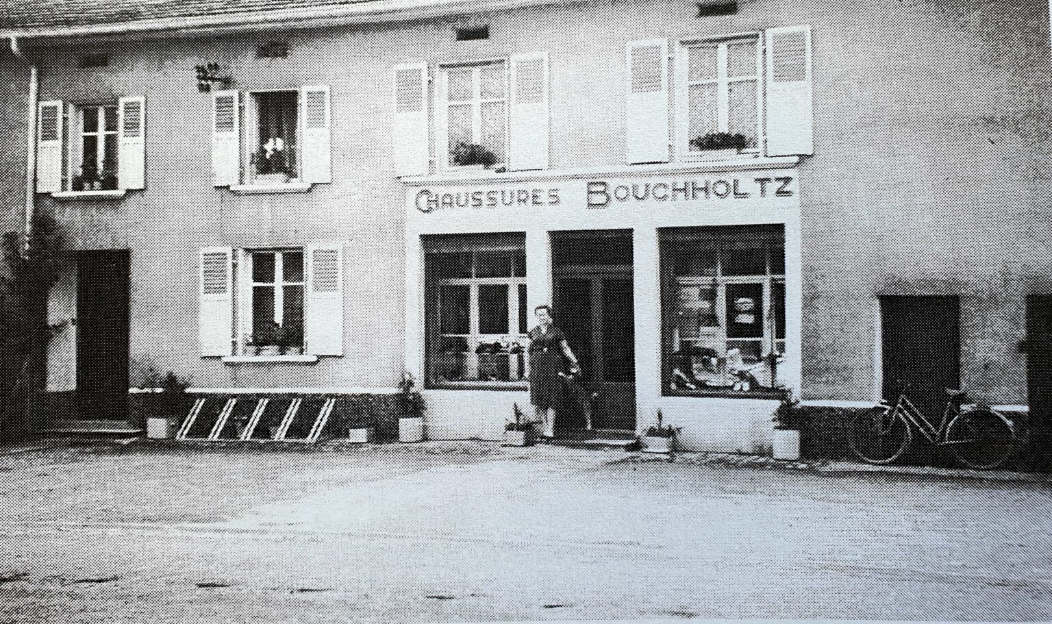 Une devanture de magasin portant l'enseigne peinte « Chaussures Bouchholtz » : deux vitrines garnies de souliers, une personne debout dans l'embrasure de la porte, et une bicyclette appuyée à droite.